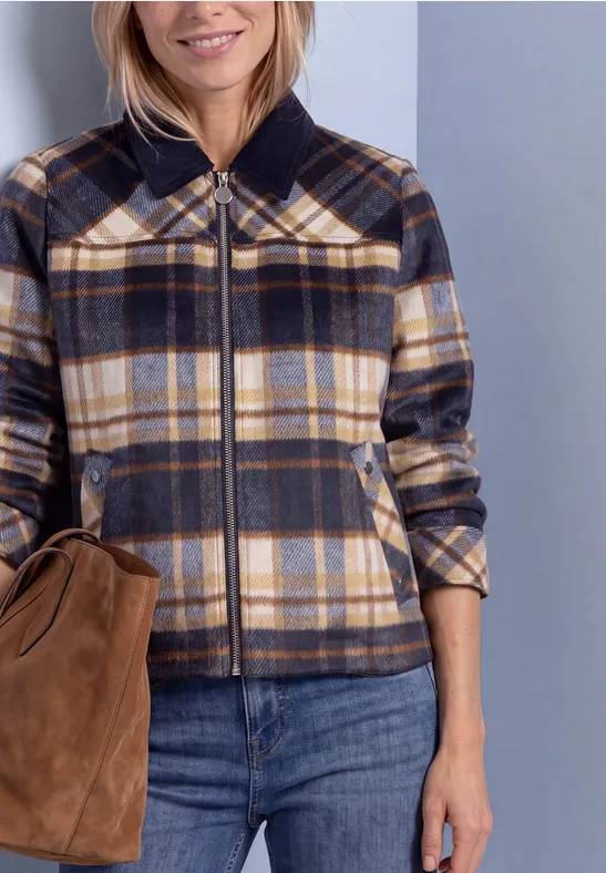 Vrouw met een kort geruit jack in neutrale kleuren, een blauwe spijkerbroek en een bruine tas.