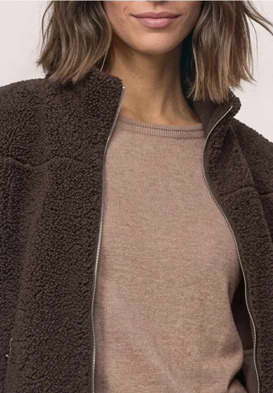 Eine Person trägt eine braune Teddy-Fleecejacke über einem hellbraunen Shirt.