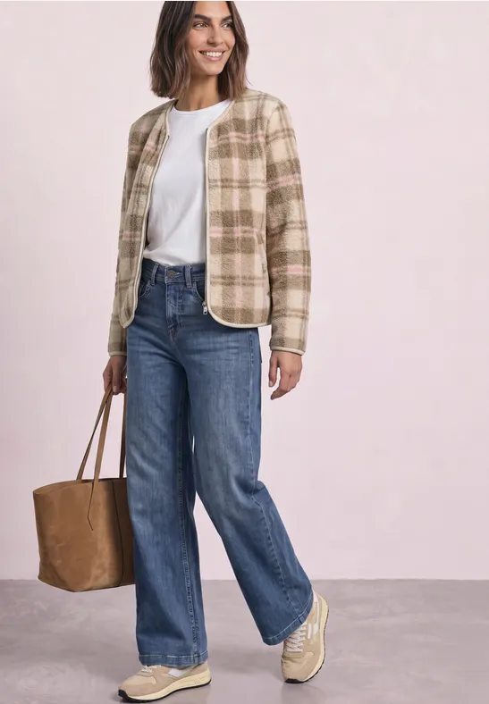 Vrouw in beige geruite jas, wit shirt, rechte jeans, beige sneakers en bruine tas voor lichte achtergrond.