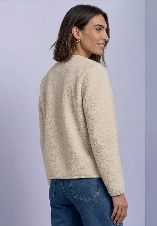 Frau in beigefarbener Fleecejacke und Jeans, von hinten seitlich im Studio vor hellem Hintergrund