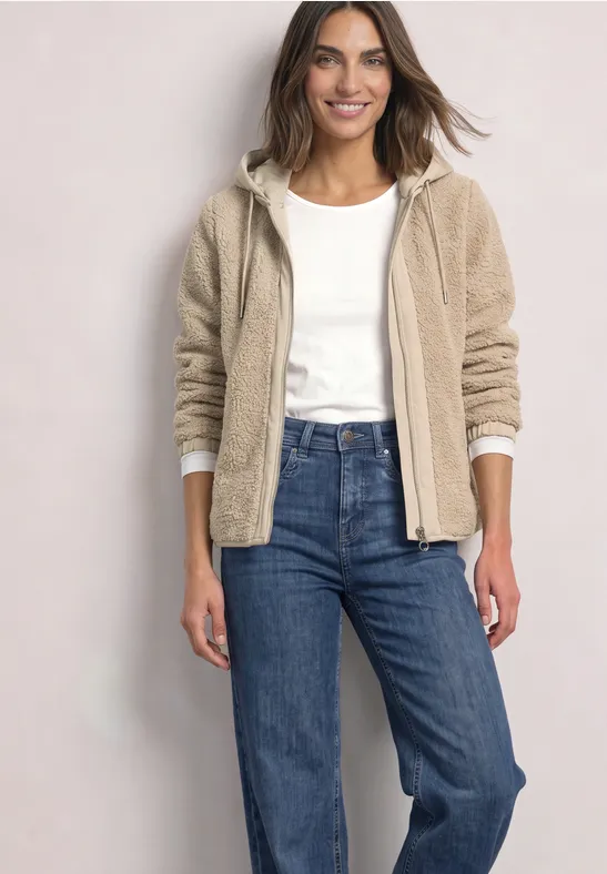 Een vrouw lacht, draagt een beige teddyjas, wit shirt en blauwe jeans.