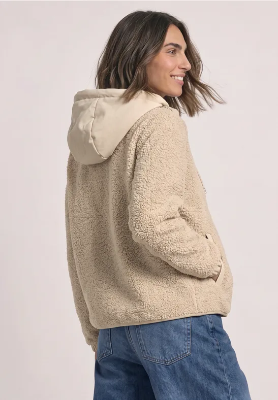 Vrouw in beige teddy-fleecejas met capuchon en blauwe jeans, glimlachend zijwaarts.