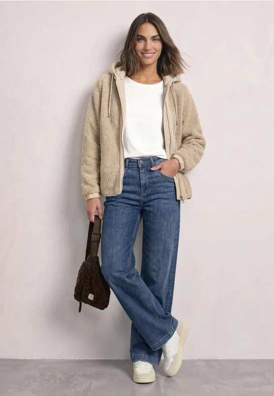 Lachende vrouw in wit topje, beige teddyjas, brede jeans, chunky sneakers en bruine tas.