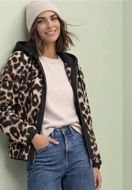 Vrouw in jeans, beige muts en luipaard-fleecejas lacht ontspannen.