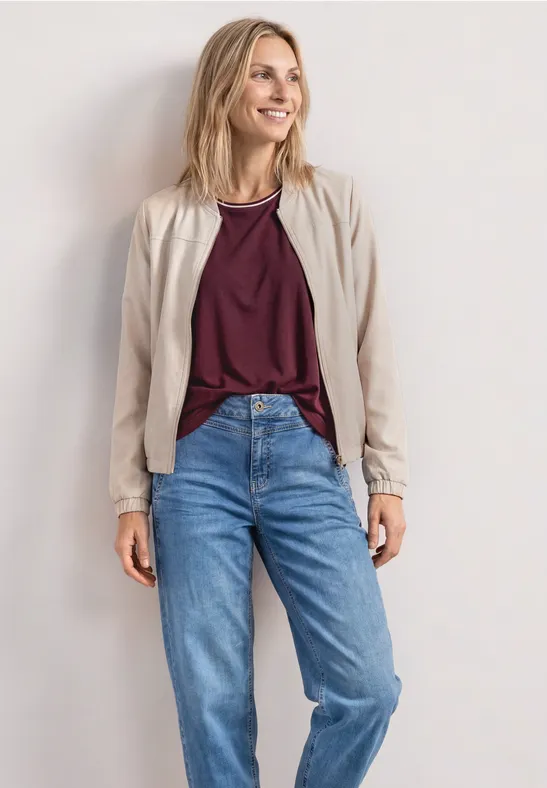 Une femme porte une veste beige, un t-shirt bordeaux et un jean bleu devant un mur blanc.