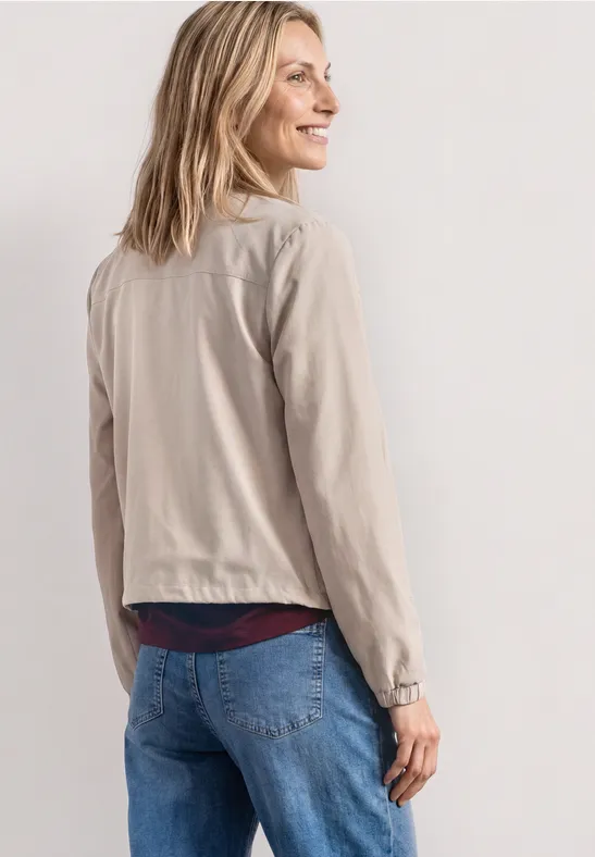 Une femme de dos porte une veste beige clair et un jean bleu devant un fond neutre.