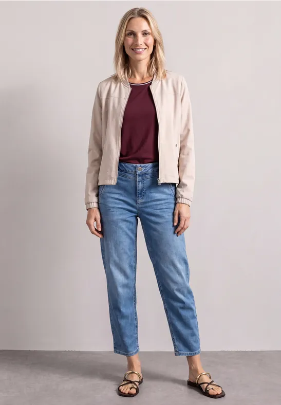 Femme portant jean, top bordeaux, veste claire et sandales noires devant un fond neutre.