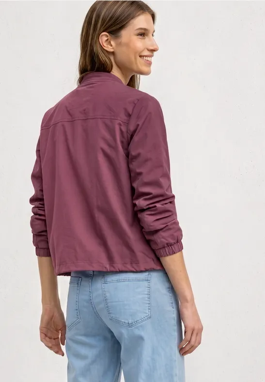 Femme de dos portant une veste bordeaux et un jean bleu clair sur fond neutre.