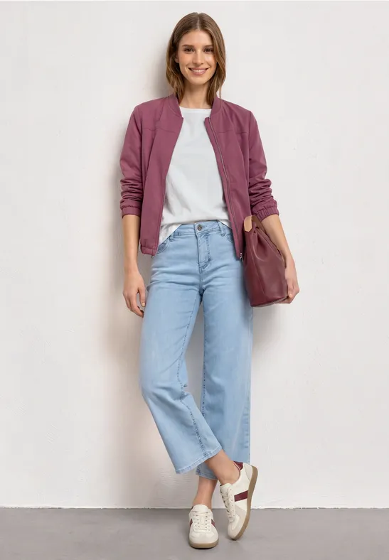 Personne porte une veste bomber violette, un t-shirt blanc, un jean clair et des baskets blanches, tenant un sac marron.