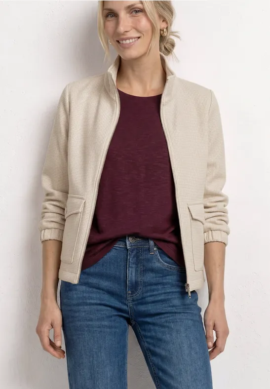 Femme devant un mur blanc, portant une veste bomber beige, un t-shirt bordeaux et un jean bleu.