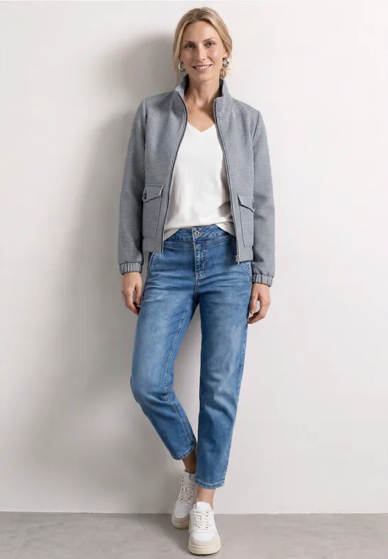 Vrouw in wit T-shirt, grijze blazer, blauwe jeans en witte sneakers voor een neutrale achtergrond.