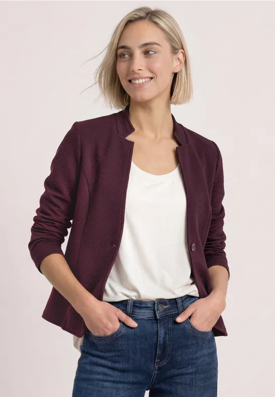 Une femme souriante en top blanc, blazer bordeaux et jeans bleu, pose les mains dans les poches.