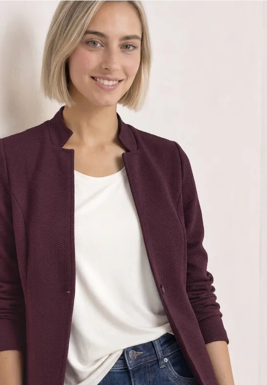 Une femme aux cheveux blonds courts souriante, en gilet bordeaux, t-shirt blanc et jean.
