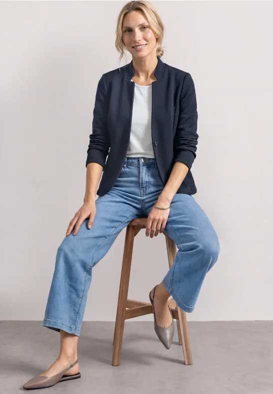 Frau sitzt auf Hocker, trägt weiße Bluse, dunkelblaue Jacke, weite Jeans und graue Ballerinas.
