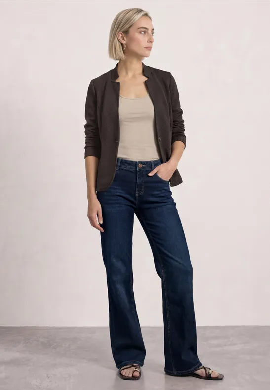 Une femme en cardigan foncé, débardeur beige, jean large bleu foncé et sandales, posant devant un fond neutre.