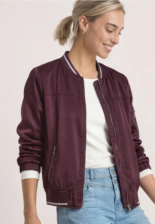 Jeune femme souriante porte une veste bomber bordeaux, un t-shirt blanc et un jean.