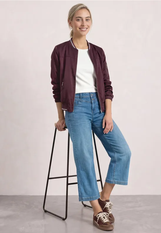 Jeune femme souriante en bomber bordeaux, t-shirt blanc et jean large appuyée sur un tabouret.
