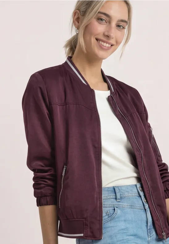 Une femme souriante porte une veste bomber violette, un t-shirt blanc et un jean bleu sur fond clair.