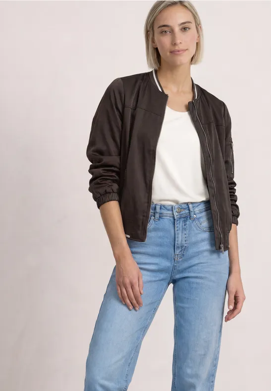 Une jeune femme porte une veste bomber foncée, un t-shirt blanc et un jean clair sur fond neutre.