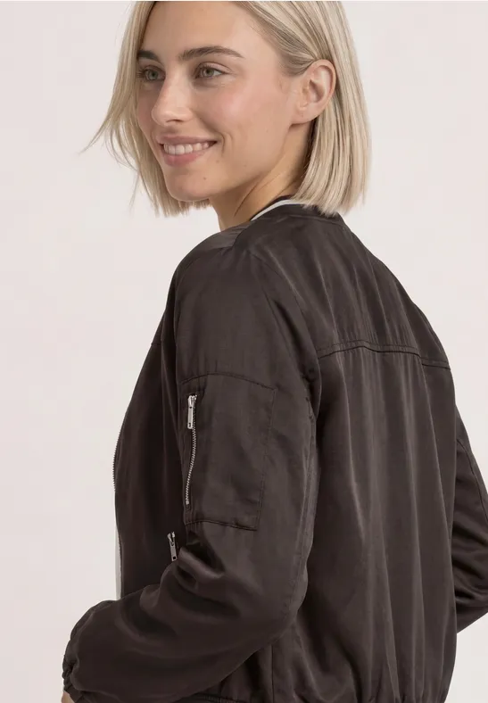 Jeune femme aux cheveux blonds courts, portant une veste bomber noire, sourit de profil.