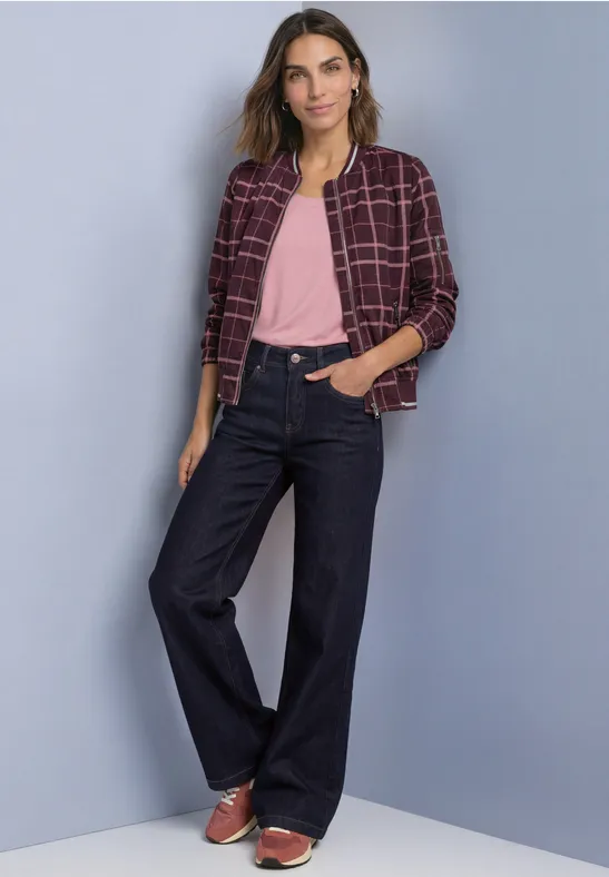 Frau in weitem dunkelblauen Jeans, rosa Shirt und kariertem Blazer steht lässig vor grauer Wand.