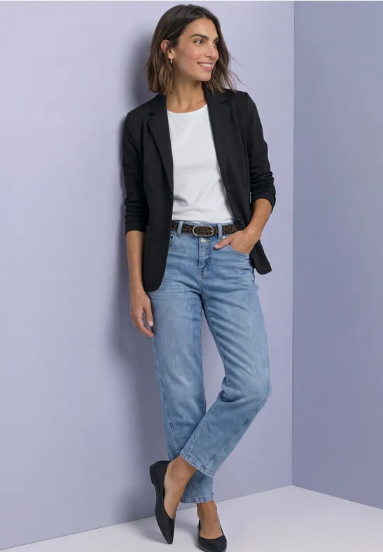 Frau mit schwarzem Blazer, weißem Shirt, Jeans und schwarzen Ballerinas vor dezent lila Wand.