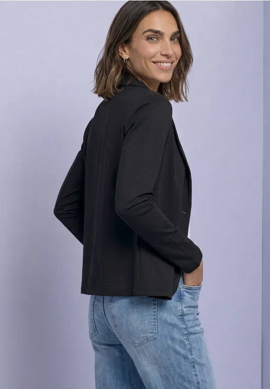 Eine Frau in schwarzem Blazer und blauen Jeans steht seitlich lächelnd vor hellem Hintergrund.