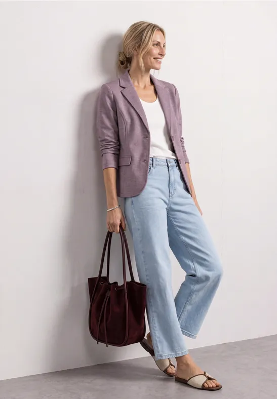 Frau in hellem Jeans-Outfit mit weißem Shirt, fliederfarbenem Blazer, Sandalen und großer dunkelroter Tasche.
