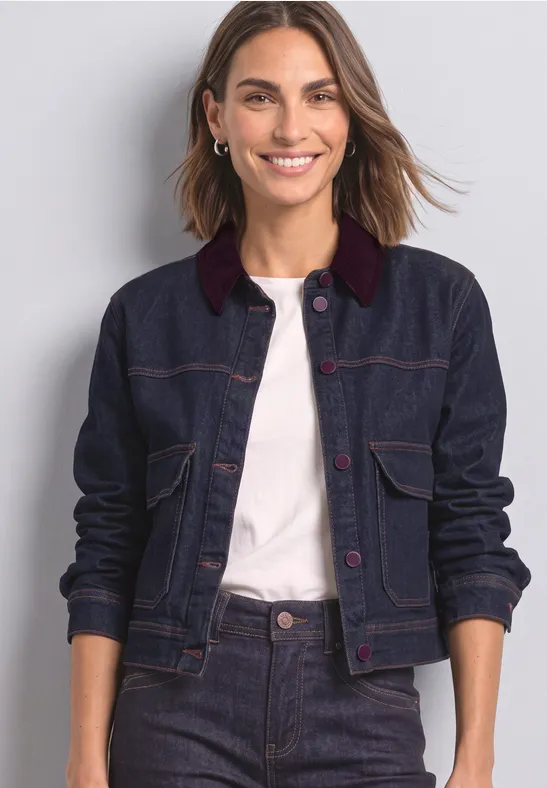 Eine lächelnde Frau trägt eine dunkelblaue Jeansjacke mit rotem Kragen und weißem Shirt vor hellgrauem Hintergrund.