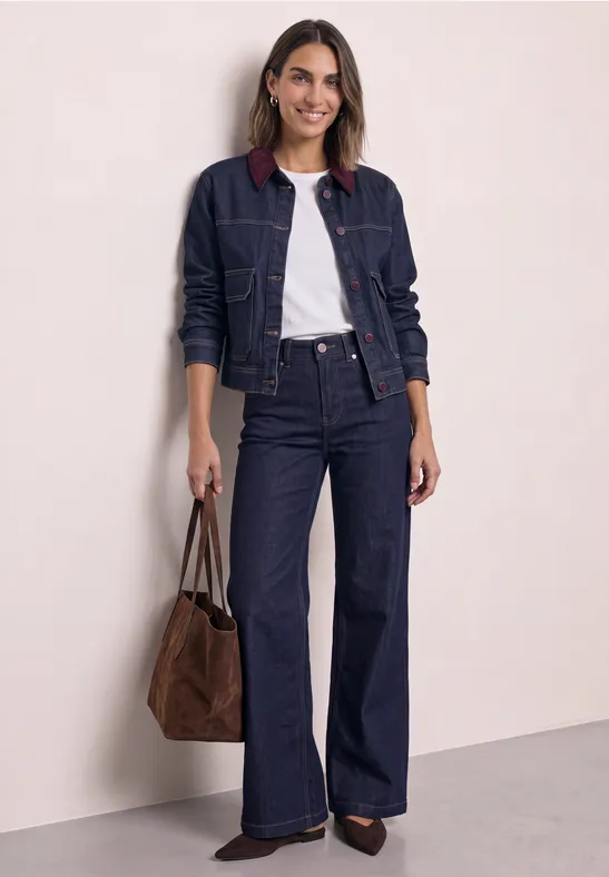 Frau in dunkelblauer Jeansjacke mit weißem Shirt, weiter Jeanshose und brauner Handtasche.