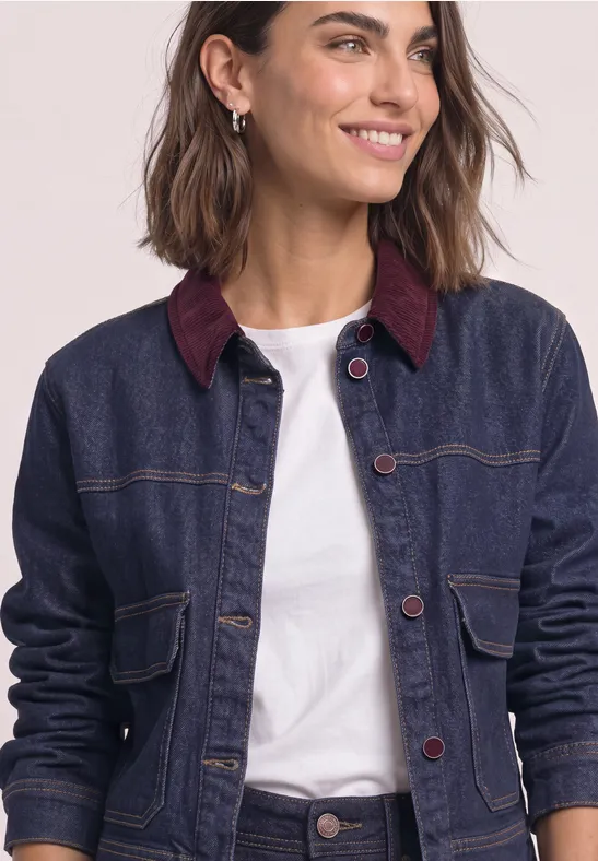 Frau in dunkelblauer Jeansjacke mit bordeauxfarbenem Kragen und weißem Shirt, lächelnd vor hellem Hintergrund.