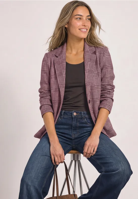 Frau im karierten Blazer, schwarzem Top und Jeans sitzt auf einem Hocker und hält eine Tasche.