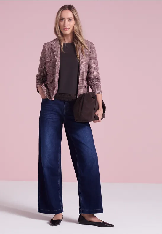 Eine Frau trägt Blazer, schwarzes Oberteil, weite Jeans und hält eine Handtasche vor rosa Hintergrund.