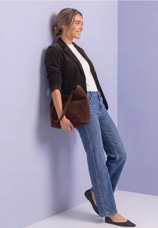 Eine Frau lehnt an einer pastellfarbenen Wand, trägt schwarze Jacke, weiße Bluse, blaue Jeans und eine braune Tasche.