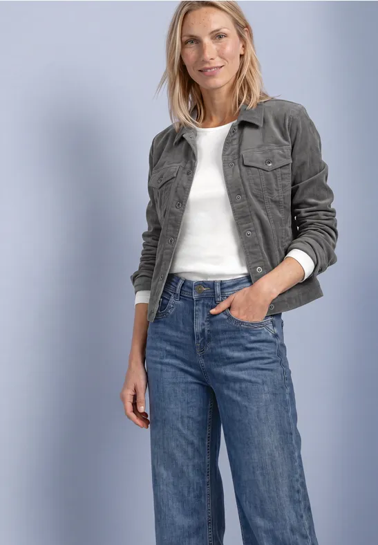 Frau in dunkelgrüner Cordjacke, weißem Top und blauen Jeans, steht lächelnd mit einer Hand in der Hosentasche.