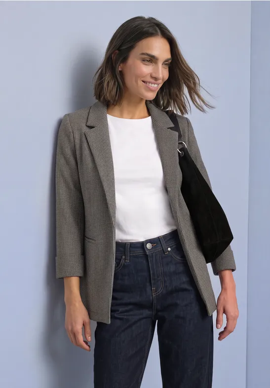 Une femme souriante porte un blazer gris, un t-shirt blanc, un jean et un sac épaule noir sur fond clair.