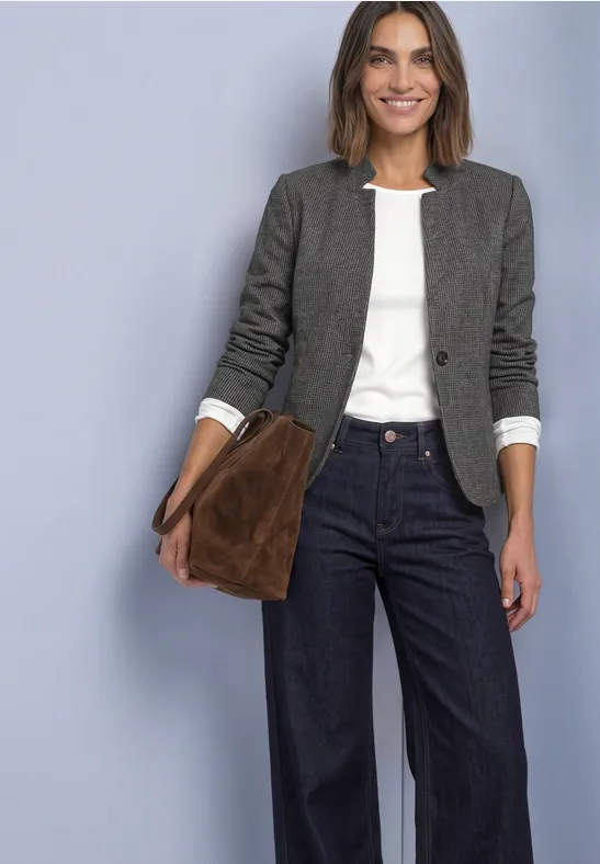 Frau in grauer Jacke, weißem Shirt, weiten Jeans und brauner Tasche vor hellblauem Hintergrund.