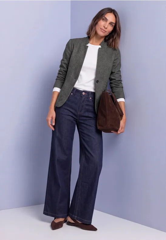 Frau in weißem Top, grüner Jacke und weiten dunkelblauen Jeans, braune Schuhe und große Clutch vor schlichtem Hintergrund.
