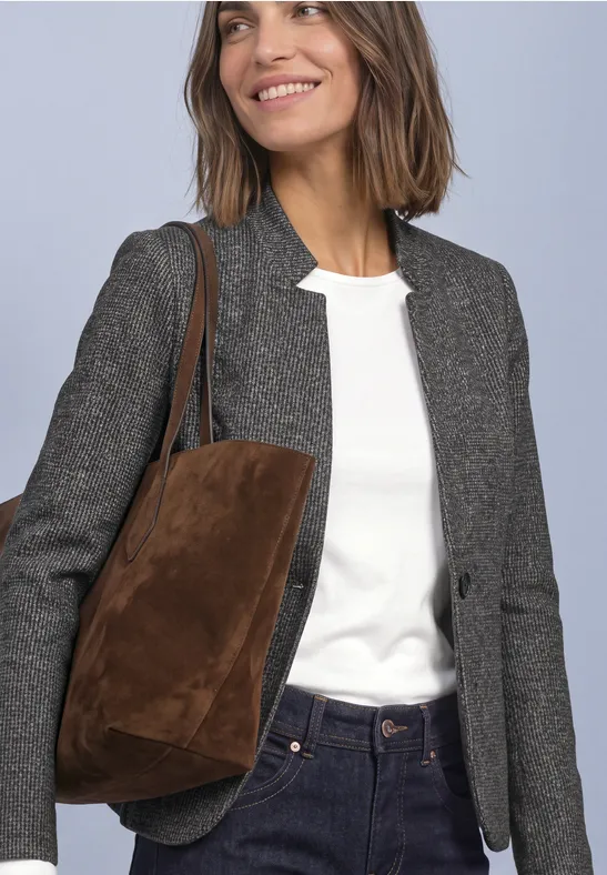 Frau mit weißem Shirt, grauem Blazer, Jeans und brauner Wildledertasche vor neutralem Hintergrund.
