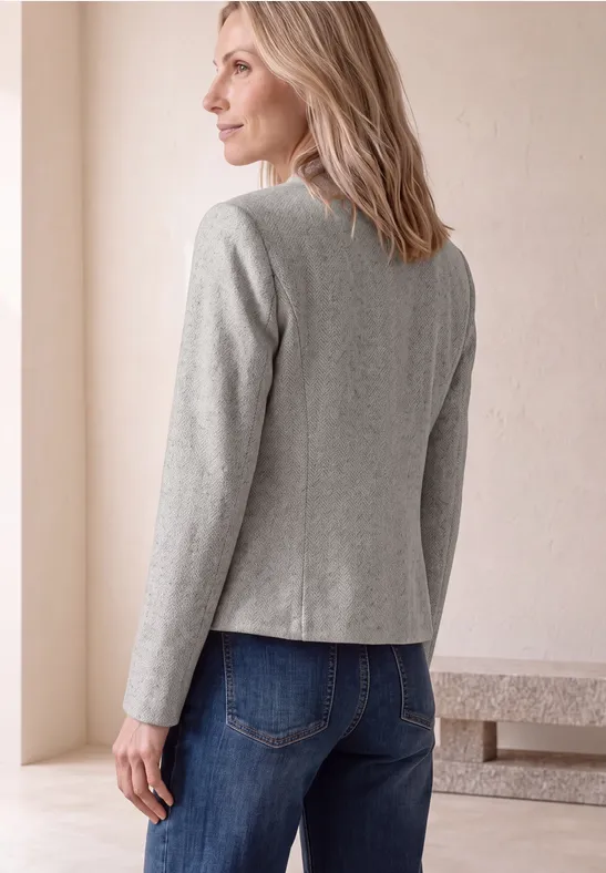 Vrouw van achteren in een grijs colbert en blauwe jeans voor een lichte achtergrond.