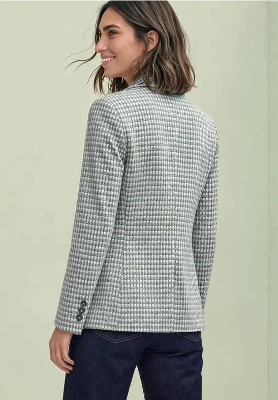 Achteraanzicht van een vrouw in een grijs pied-de-poule jasje en donkere jeans tegen lichte achtergrond.