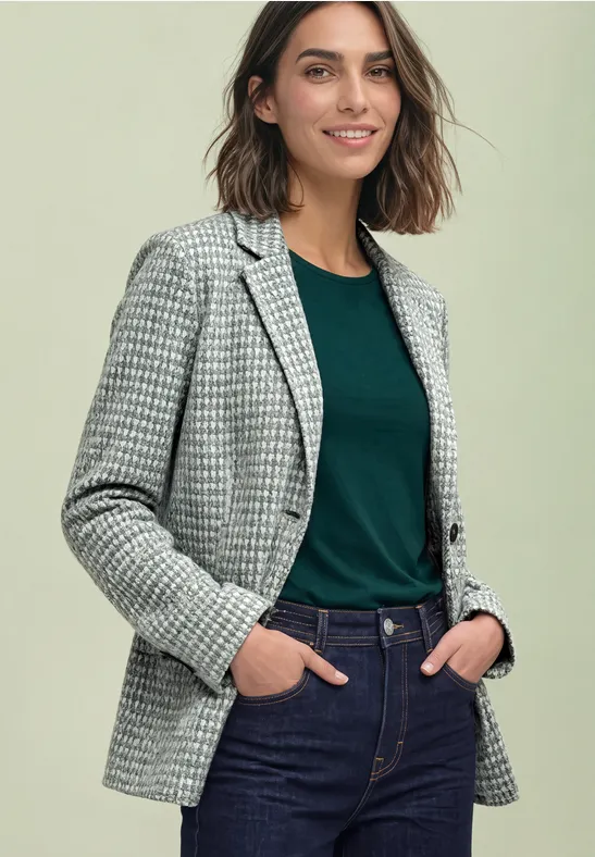 Vrouw met pied-de-poule blazer, groen shirt en spijkerbroek, handen in zakken, glimlachend.