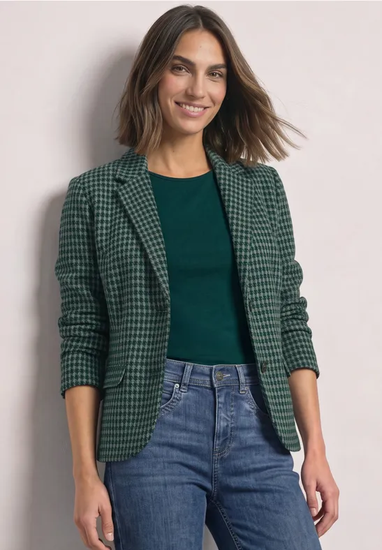 Femme souriante en veste à carreaux verts, top vert et jean devant un fond clair