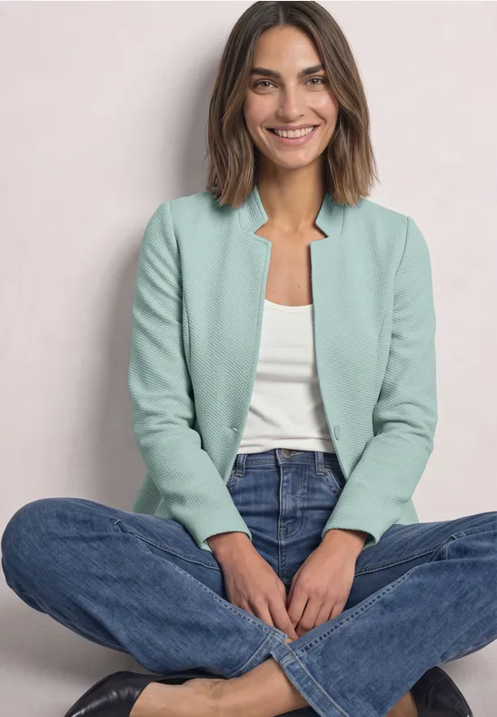 Une femme souriante est assise jambes croisées, portant un blazer vert clair, un haut blanc et un jean bleu.