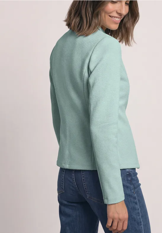 Femme de profil portant un blazer vert pâle et un jean, souriante sur fond neutre.