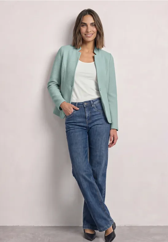Femme en veste vert menthe, débardeur blanc et jean bleu sur fond clair.