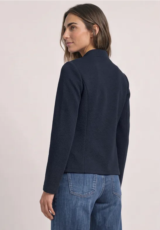 Eine Frau von hinten blickt zur Seite, sie trägt ein dunkelblaues Sakko und blaue Jeans.