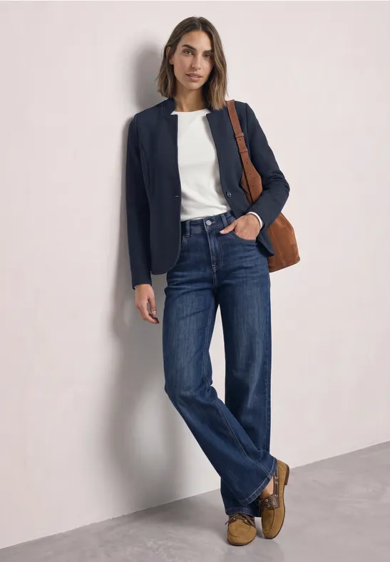 Frau in weißem Shirt, dunkelblauem Blazer, geraden Jeans, braunen Mokassins und Schultertasche vor hellem Hintergrund.
