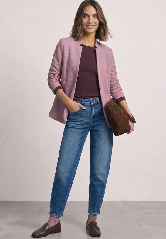 Une femme porte un blazer rose, un t-shirt bordeaux, un jean bleu et des mocassins, tenant une pochette marron.