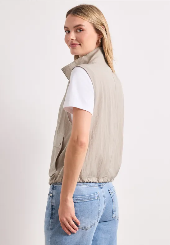 Seitlich stehende Person trägt beiges ärmelloses Jackett über weißem T-Shirt und Jeans vor weißem Hintergrund.